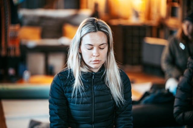 A woman sitting on the ground wearing a black jacket and breathing through her nose.