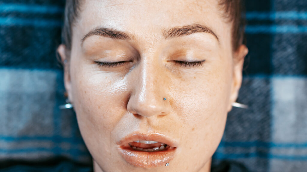 Close-up of a person experiencing holotropic breathwork, with eyes closed and a serene expression. The individual’s face, showcasing natural skin texture and facial piercings, conveys a sense of deep relaxation and emotional release.