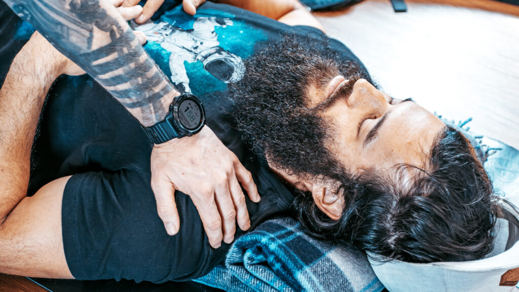 A man laying down practising Holotropic breathwork while another man puts his hand on his shoulder.